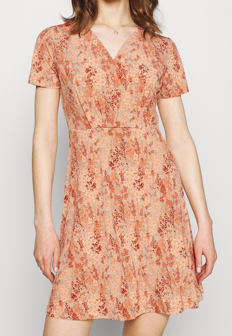Kwiecista sukienka typu wrap w brzoskwiniowo-beżowym kolorze z krótkimi rękawami. Posiada dekolt w kształcie litery V oraz delikatne czerwone wzory kwiatowe w całej sukience. Gładka faktura.