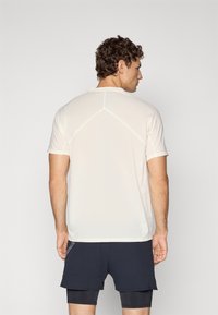 Man facing away wears light beige short-sleeve sports shirt and black layered running shorts against plain background.