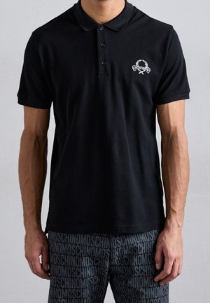 Homme portant un polo noir à manches courtes avec un petit logo blanc et un short foncé avec un motif de texte répété.