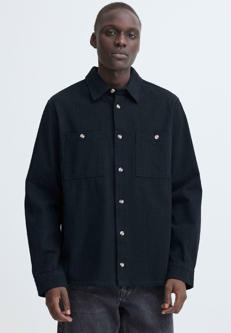 Άνδρας που φοράει μαύρο πουκάμισο με κουμπιά και δύο τσέπες στο στήθος, καθώς και σκούρα τζιν, στέκεται μπροστά σε ένα απλό λευκό φόντο.