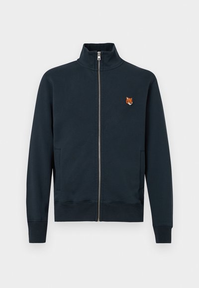 Giacca zip-up blu navy con collo alto, dotata di tasche laterali e un piccolo logo di una volpe ricamato sul petto, realizzata in materiale morbido.