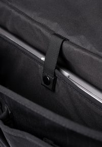 Interno di una borsa per laptop in tessuto nero con un comparto imbottito, una cinghia sicura e un bottone a pressione, adatta per proteggere i dispositivi.