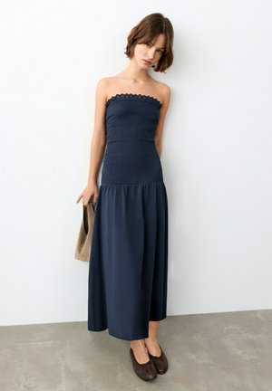 Jeune femme portant une robe bleu marine sans bretelles avec un haut à volants, tenant un sac à main beige, debout sur un sol gris devant un mur blanc.