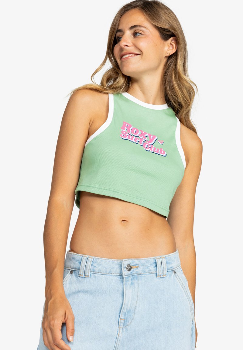 Top verde con ribete blanco y gráfico rosa "ROXY Surf Club". Hecho de algodón con un diseño ajustado. Lleva puesto con shorts de mezclilla azul claro.