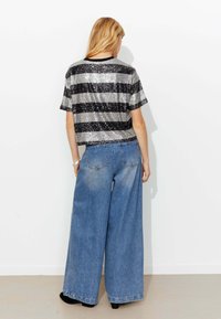 Chemise courte rayée noire et argentée à sequins, associée à un jean denim bleu clair à jambes larges. La chemise a des manches courtes et un col rond.