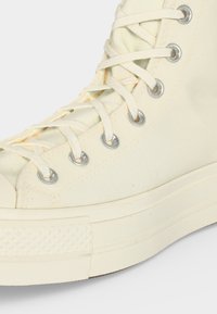 Λευκό sneaker καμβά ψηλής κορυφής με λευκά κορδόνια περασμένα από μεταλλικές τρύπες και παχιά λαστιχένια σόλα.