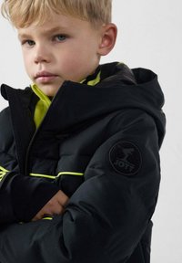 Chaqueta acolchada negra con detalles en amarillo neón y un parche con logo en la manga. Con capucha, cremallera frontal y puños elásticos.