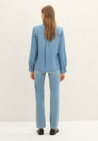 Camisa de mezclilla azul claro con mangas largas, puños abotonados y dobladillo curvado, combinada con unos pantalones vaqueros azul claro a juego y botas negras de tobillo.
