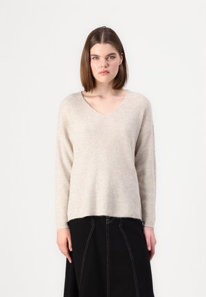 Beige stickad tröja med v-ringning och långa ärmar. Mjuk textur, avslappnad passform, slutar vid höfterna. Matchas med en svart kjol.