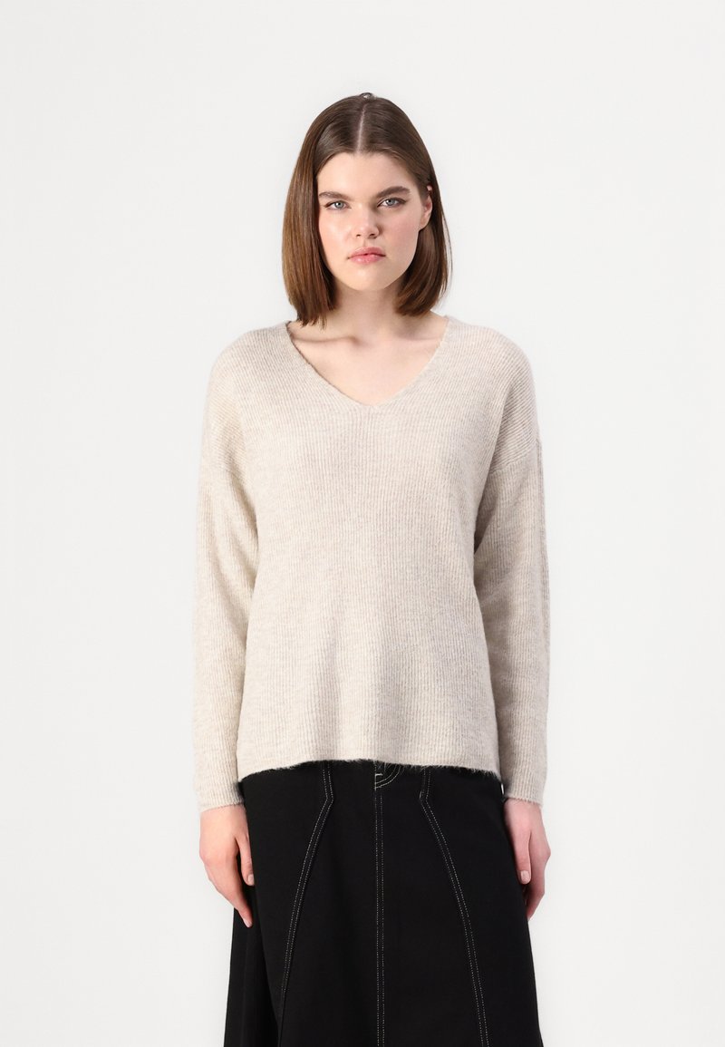 Maglione beige lavorato a maglia con scollatura a V e maniche lunghe. Texture morbida, vestibilità rilassata, che arriva ai fianchi. Abbinate a una gonna nera.