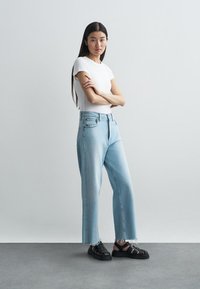 Lyseblå høytlivsjeans med rett kutt, utslitt nederkant, sammen med en hvit tettsittende t-skjorte og svarte sandaler med stropper.