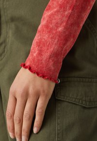Rode lange mouwen top met een gestructureerde afwerking en gerimpelde manchetten, gedragen over een olijfgroen kledingstuk. Hand rustend op de stof.