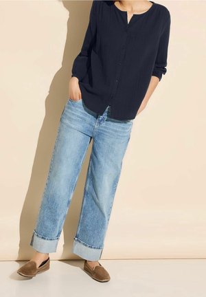 Person iført en løs sort skjorte med knapper, lyseblå vide bukser med manchetter og brune slip-on sko, står mod en beige baggrund.