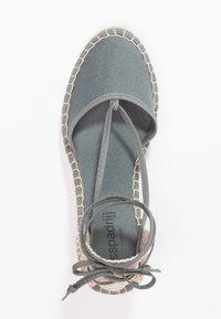 Šedé plátěné espadrilky s kulatou špičkou, tkanou jutu podrážkou a zkříženými šňůrkami kolem kotníku. Podešev obsahuje kontrastní švy.