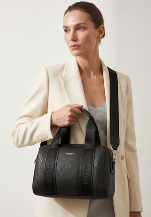 Vrouw in crème blazer houdt zwarte Diana&Co leren handtas met gebrande banden over haar schouder tegen een effen achtergrond.