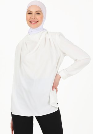 White long-sleeve blouse with draped front detail, soft fabric, rounded hem, and cuffed sleeves, styled with black pants.