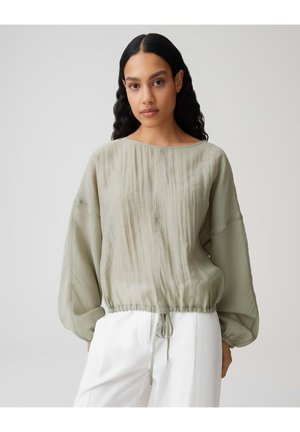 Femme aux longs cheveux noirs portant un chemisier transparent vert clair à manches longues avec cordon de serrage à l'ourlet et un pantalon blanc ample, debout devant un fond uni.