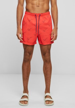 Shorts de bain rouges avec un motif de homard, taille élastique, cordon bleu ajustable et coupe mi-cuisse. Portés avec des sandales beige.