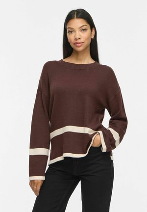Femme aux longs cheveux foncés portant un pull marron à rayures blanches et un pantalon noir, debout avec la main dans la poche sur un fond uni.
