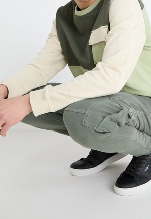 Person hockt, trägt einen farblich abgesetzten grünen und beigen Sweatshirt, grüne Cargohosen und schwarze Sneakers mit weißen Sohlen.