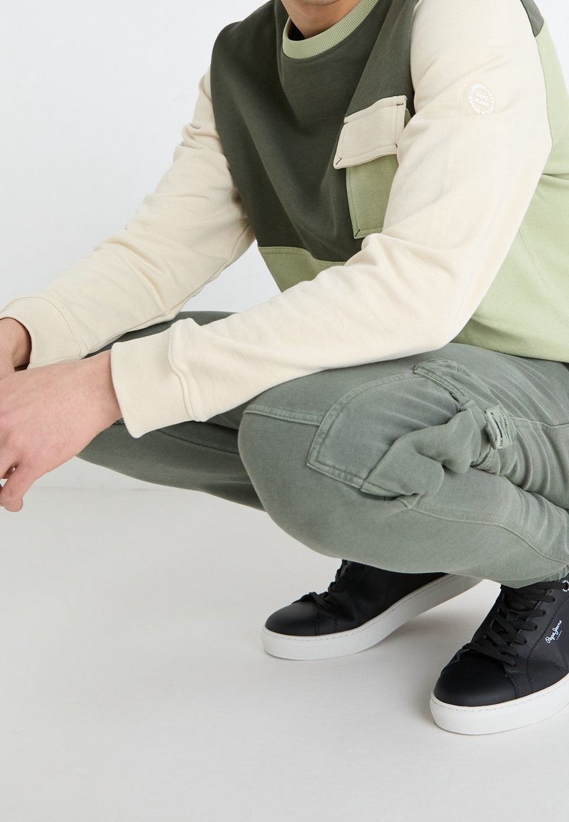 Personne accroupie, vêtue d’un sweat-shirt bicolore vert et beige, d’un pantalon cargo vert et de baskets noires à semelles blanches.