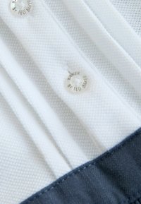 Biały materiał o fakturze z głębokim niebieskim akcentem; zdobiony srebrnymi guzikami z napisem "BAKER BY TED BAKER." Wzór zawiera pionowe plisy.