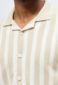 Κοντινό πλάνο ενός ατόμου που φοράει μπεζ και λευκό κάθετα ριγέ πλεκτό πουκάμισο με γιακά και τρία λευκά κουμπιά.