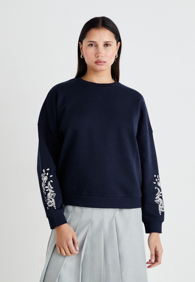Sudadera azul marino con mangas largas y relajadas, que presenta bordados florales blancos en una de las mangas. Material suave con escote redondo.