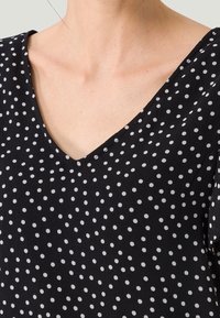 Top nero a V con pois bianchi. Tessuto leggero e testurizzato. Maniche corte che terminano sulla spalla. Design semplice e versatile.