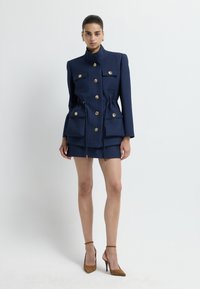 Donkerblauwe jurk met een textuurstof, gouden knopen, twee zijzakken, een getailleerde taille en een korte aansluitende rok. Bruine hoge hakken.