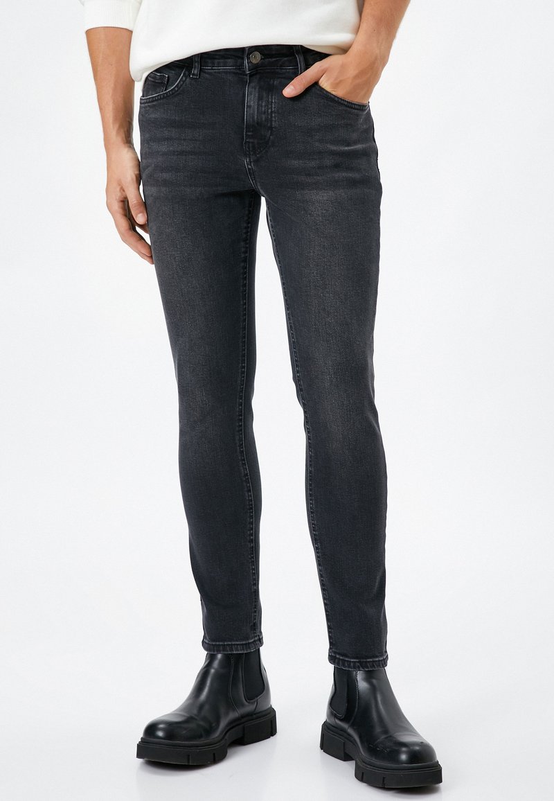 Jeans negros ajustados para hombre, hechos de denim, con bolsillos frontales, pasadores para cinturón y un ligero desgaste, combinados con botas negras hasta el tobillo.