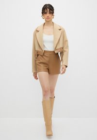 Beige cropped jakke, hvid tætsiddende top, brune lædershorts og knæhøje sandfarvede støvler. Jakken har store klapper og hardware detaljer på siden.