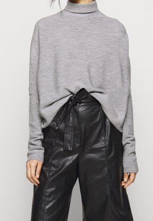Femme portant un pull à col roulé gris rentré dans un pantalon en cuir noir taille haute avec une ceinture nouée à la taille.