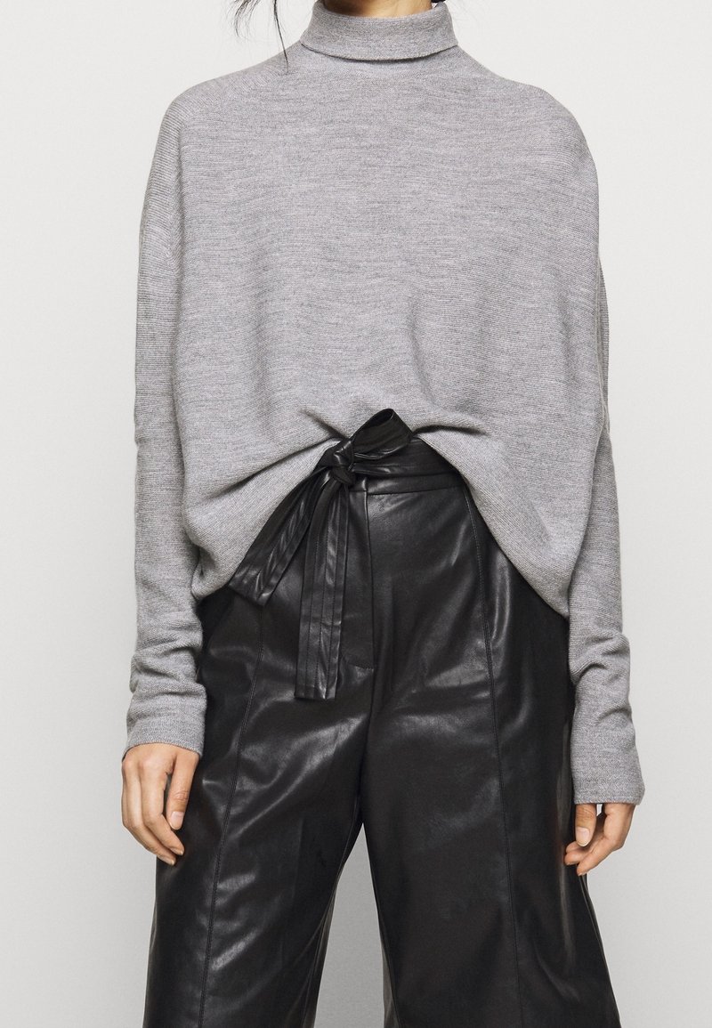 Femme portant un pull à col roulé gris rentré dans un pantalon en cuir noir taille haute avec une ceinture nouée à la taille.