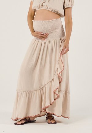 Frau in beigem gerafftem Crop-Top und passendem langem Rock mit Rüsche, hält ihren Schwangerschaftsbauch, trägt dunkle Sandalen.