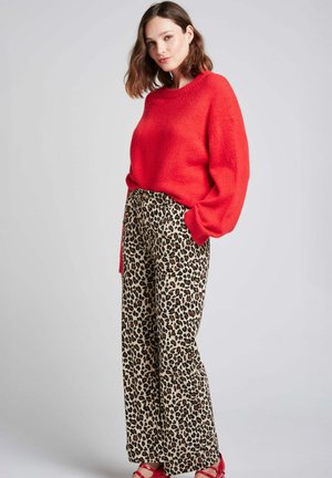 Pull rouge texturé avec épaules tombantes, associé à un pantalon large en imprimé léopard avec un fond crème et des taches marron.