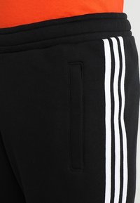 Zwarte sweatpants met witte strepen langs de zijkant, een elastische tailleband en een zijzak. De stof is zacht met een gladde textuur.