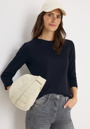 Chemise à manches longues bleu marine avec un col rond, associée à une casquette beige clair et un pantalon gris. La casquette a une texture douce et des coutures détaillées.