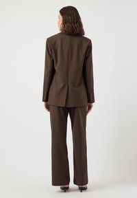 Completo marrone su misura con blazer strutturato, polsini con bottoni e pantaloni dritti, realizzato in tessuto liscio e intrecciato.