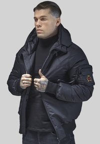 Schwarze Daunenjacke mit hohem Kragen, Reißverschlusstaschen und gerippten Bündchen. Das Material wirkt glatt und hat ein gesticktes Patch am Ärmel.