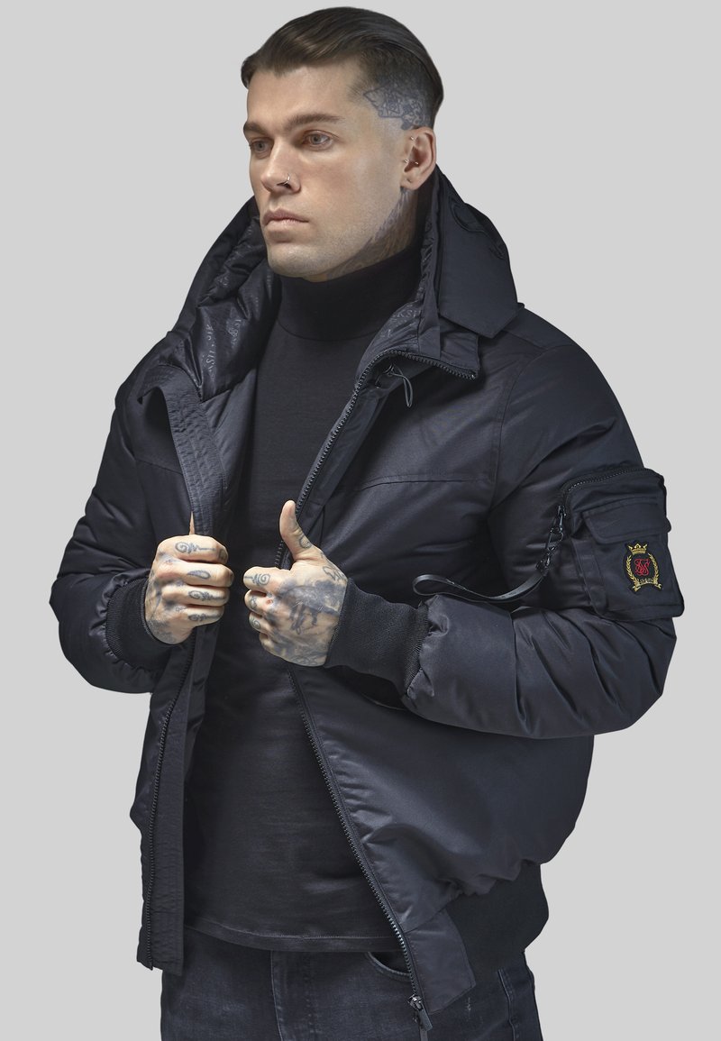 Schwarze Daunenjacke mit hohem Kragen, Reißverschlusstaschen und gerippten Bündchen. Das Material wirkt glatt und hat ein gesticktes Patch am Ärmel.