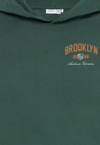 Zelená mikina s kapucňou, na ktorej je nápis "BROOKLYN 1986" v oranžovej a bielej farbe. Mäkká textura, klasický strih a kapucňa so šnúrkou.