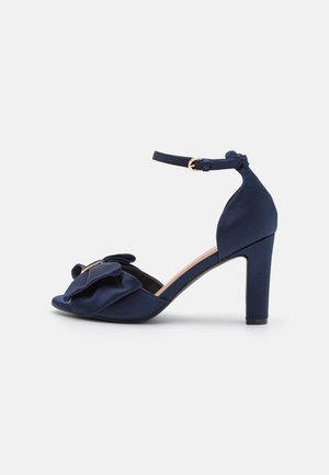 Marineblaue Satin-Sandale mit hohem Absatz, Knöchelriemen, Schnalle und dekorativer Schleife am offenen Zehenbereich.