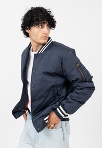 Marineblauwe bomberjack met witte gestreepte geribbelde kraag en boorden, ritszak op de linker mouw, gladde stof en knoopsluiting aan de voorkant.