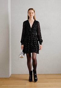 Abito nero con design avvolgente, maniche lunghe e motivo a pois bianchi. Abbinato a una borsa argento e stivaletti alla caviglia, su uno sfondo neutro.