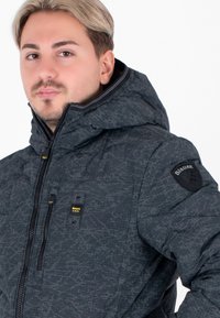 Giacca a piumino nera con motivo e colletto alto, dotata di cappuccio. Presenta cerniere argento e una toppa circolare con logo sulla manica sinistra.