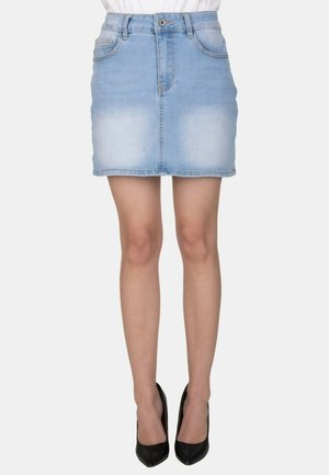 Minigonna in denim azzurro chiaro indossata con tacchi alti neri su una persona in piedi davanti a uno sfondo semplice.