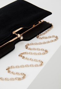 Clutch in suede nera con una chiusura in metallo dorato, caratterizzata da una pietra decorativa. La tracolla in catena dorata è posizionata lungo la borsa, con segmenti collegati.