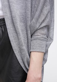 Maglione grigio a maglia con vestibilità ampia e spalle scese. Presenta polsini arrotolati e una texture morbida. Abbinato a una gonna in pelle nera.