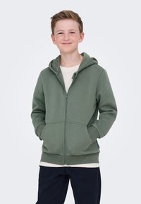 Niño sonriente con cabello corto, vestido con una sudadera verde salvia con cremallera, una camiseta blanca y pantalones negros, con las manos en los bolsillos, de pie sobre un fondo blanco.